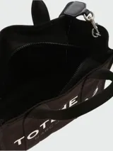 Bolso tipo tote de lona color negro con estampado frontal en blanco que dice 'THE TOTE BAG' y 'MARC JACOBS'. Cuenta con doble asa superior, correa ajustable y extraíble para llevar al hombro y cierre superior con cremallera.