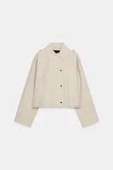 Trench corta de color beige, con cuello solapa, mangas largas con trabillas en los hombros, bolsillos delanteros de vivo y cierre frontal cruzado con botones a presión.