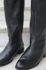 Bota de caña alta confeccionada en cuero, con diseño de textura perforada en la parte superior de la caña y puntera lisa. Cuenta con suela plana y taco bajo.
