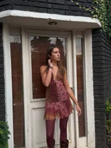 Vestido corto negro con escote en V profundo, estilo halter con tiras para atar al cuello. Presenta un diseño ajustado en el busto con tiras ajustables bajo el mismo que se cruzan y anudan en la espalda, y falda con volado en el ruedo con detalle de encaje.