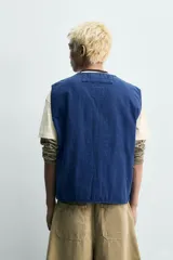 Chaleco de algodón azul, de corte relaxed fit, ligeramente acolchado con pespuntes, cuello en pico, cierre frontal con cremallera y bolsillos de plastrón en la cadera.