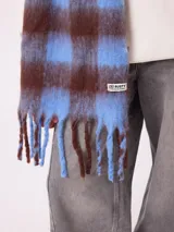 Bufanda de tejido suave con diseño de cuadros grandes en tonos naranja, rojo, violeta y lila. Presenta flecos en los extremos y una etiqueta pequeña con el logo de la marca Rusty.