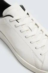 Zapatilla de cordones confeccionada en piel, con horma redonda y suela a tono. Presenta un diseño minimalista con detalle de contraste en el borde del talón.