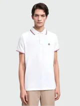 Polo de piqué de algodón en color blanco, con cuello y puños acanalados que presentan detalles de rayas tricolor. Cuenta con cierre frontal de botones y el logo de la marca aplicado en el pecho.
