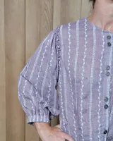 Camisa de manga larga con diseño de rayas verticales y bordados sutiles. Presenta cuello redondo, cierre frontal con botones y mangas abullonadas con volados en los hombros.
