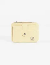 Tarjetero/monedero compacto de cuero vegano color amarillo pálido, con cierre a presión en solapa frontal y bolsillo superior con cierre de cremallera. Presenta múltiples ranuras para tarjetas y un dije metálico con el número "5" en la parte inferior derecha.