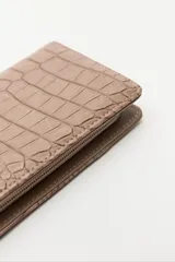 Billetera de mano con textura efecto cocodrilo, color beige, con cierre perimetral y tira de muñeca desmontable.