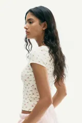 Remera corta de punto acanalado color crema, con estampado floral diminuto en tonos rosados y marrones. Presenta escote cuadrado con ribete de picot y mangas cortas ajustadas.