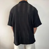 Camisa de tejido calado con textura, color blanco, de corte oversize, manga corta y cuello solapa con cierre frontal de botones.