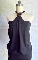 Vestido corto negro, sin mangas, con escote halter que se ajusta con una argolla oscura en el centro del pecho. La parte superior tiene un drapeado suelto y la falda es ajustada.