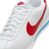 Championes Nike modelo Cortez, de diseño clásico en color blanco con el logo de la marca en rojo vibrante en los laterales y talón. Presenta una entresuela con una franja azul distintiva y suela dentada para mayor tracción.