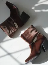 Bota corta de velvet azul marino con efecto abullonado en el tobillo y cierre lateral con cremallera. Tiene un tacón cuadrado y una cadena desmontable con el logo de la marca.