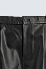 Bermuda de corte relajado confeccionada en piel negra, con pliegues frontales, bolsillos laterales y cierre frontal con cremallera y botón.