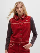 Chaleco de pana rojo con cuello camisero, cierre frontal con botones metálicos y detalles de líneas claras en cuello, canesú y ruedo. Cuenta con un pequeño parche con la letra 'M' en el pecho.