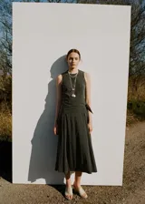 Vestido midi evasé color verde oscuro, sin mangas con tirantes anchos, cuello redondo y detalle de pañuelo cruzado en la cintura con lazo lateral. Cuenta con cierre de cremallera lateral.