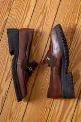 Mocasín de cuero vacuno color marrón, con suela track de goma negra, puntera cuadrada y detalle de herraje metálico sobre una banda de gamuza en el empeine.