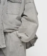 Camisa oversize de algodón orgánico con certificación GOTS, color gris claro con efecto lavado. Presenta cuello, cierre frontal de botones, dos bolsillos de parche en el pecho y puños elásticos.