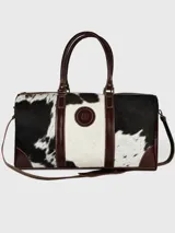 Bolso de viaje tipo duffel de cuero marrón con paneles centrales de pelo de vaca en blanco y negro. Cuenta con asas de mano y correa ajustable para hombro, ambos en cuero marrón. Presenta un parche circular con un logo grabado en el centro.