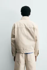 Cazadora de algodón color beige claro, corte relajado, con cuello subido y manga larga. Cierre frontal con cremallera oculta por solapa con cierres metálicos tipo alamar.