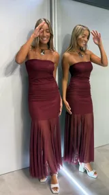 Vestido largo color bordó, de estilo strapless con escote recto. Presenta un cuerpo ajustado con detalles de fruncido en la zona de la cintura y cadera, y una falda inferior confeccionada en tela semitransparente con diseño plisado.