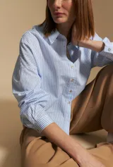 Camisa a rayas verticales azules y blancas, de manga larga con puños abotonados y cuello camisero. Presenta cartera frontal con botones dorados y un calce relajado.