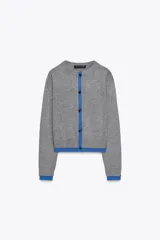 Chaqueta de punto gris de manga larga, cuello redondo y cierre frontal con botones oscuros. Presenta ribetes y detalles en color azul eléctrico en el frente, puños y ruedo.