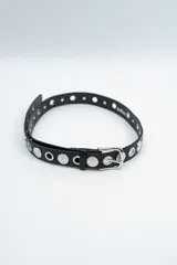 Cinturón negro con ojales metálicos y tachas circulares plateadas con diseño grabado. Cuenta con hebilla metálica plateada.