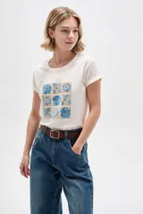 Remera de manga corta con cuello redondo y diseño holgado, en color blanco con estampado floral en tonos azules y destellos dorados.