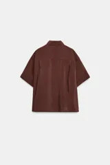 Camisa de cuello solapa y manga corta con dobladillo. Presenta un diseño de corte holgado con bajo asimétrico y aberturas laterales. Cierre frontal mediante botones ocultos bajo una tapeta.