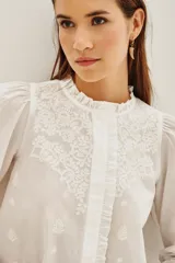 Camisa blanca de algodón con cuello alto, cierre frontal de botones ocultos y detalles de bordado floral en el pecho. Presenta mangas largas con puños elásticos y volados decorativos en la parte frontal.