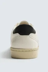 Zapatilla deportiva de estilo retro, color blanco con detalles en gris y negro. Presenta una suela de color beige en contraste y cordones blancos.