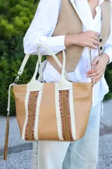 Cartera tipo shopper confeccionada en cuero vacuno con combinación de tonos nuez, camel y arena. Presenta una textura grabada, detalles en cuero liso y gamuza, doble asa de mano y correa larga ajustable. Incluye un colgante de flecos lateral y cierre de mosquetón.