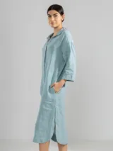 Camisa larga celeste confeccionada en lino, con cuello clásico, mangas 3/4, silueta fluida, dobladillo curvado en las costuras laterales y bolsillos discretos.