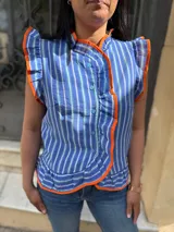 Blusa sin mangas a rayas azules, blancas y verde agua, con cuello alto y cierre frontal de botones. Destaca por sus volados ribeteados en color naranja en el cuello, hombros y la botonera.