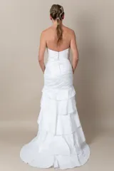 Vestido de novia blanco, sin mangas, con escote corazón y espalda en V. Confeccionado en tafeta drapeada con bordados blancos. Falda con volados y tajo frontal.