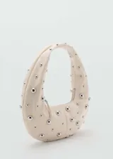 Bolso de hombro con diseño ovalado tipo hobo, confeccionado en material efecto piel color beige, decorado con tachuelas metálicas plateadas distribuidas por toda la superficie y cierre de cremallera.