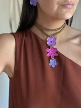 Collar tipo choker con tiento de gamuza trenzado y dije colgante compuesto por tres flores metálicas pintadas a mano en tonos violeta y fucsia, con detalles de mostacillas y un cristal central.