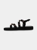 Sandalias planas de cuero gamuza en color negro, con múltiples tiras cruzadas decoradas con tachas metálicas doradas y aplicaciones de piedras rojizas. Cuenta con suela de PVC y tira ajustable en el talón con hebilla metálica dorada.