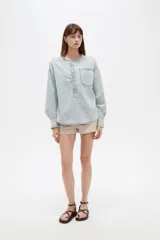 Blusa de jean ligera en color celeste claro, con cuello redondo y cierre frontal parcial con botones. Presenta un diseño con volados a lo largo de la botonera, bolsillo ojal en el pecho, y mangas largas abullonadas con puños ajustados y fruncidos.