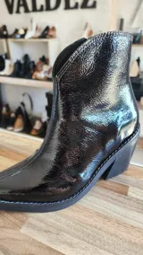 Botas tejanas de cuero charolado negro, con taco cuadrado de 7 cm y punta fina. Cuentan con cierre interno y costuras visibles en la suela.