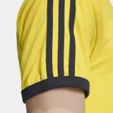 Remera de algodón Adidas Adicolor Sport de color amarillo, con cuello redondo y ribetes negros. Cuenta con las icónicas tres tiras negras en las mangas y el logo del trébol bordado en el pecho.