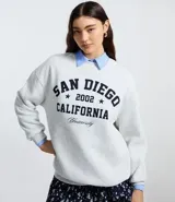 Suéter de cuello redondo con diseño estilo college, color gris melange con estampado frontal en letras oscuras. Presenta puños y ruedo acanalados.