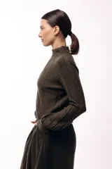 Polera de tejido de punto color marrón, con cuello alto y diseño de trenzas verticales. Presenta mangas largas con puños ajustados y un detalle de panel lateral asimétrico que se anuda en la cintura.