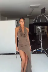 Vestido midi de color marrón, con diseño de cuello halter y escote drapeado. Presenta un corte ajustado con frunces laterales y una abertura pronunciada en la pierna que le otorga un estilo asimétrico.