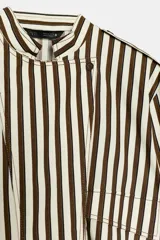 Camisa tipo bomber confeccionada en algodón, con diseño de rayas verticales en tonos marrón y blanco. Presenta cuello perkins, manga larga con puños, bolsillo de plastrón con solapa en el frente, bajo elástico y cierre frontal con botones.
