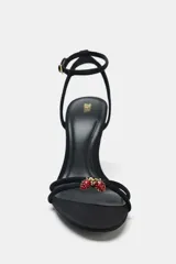Sandalia de tacón alto fino, color negro, con tiras finas cruzadas en el empeine y un detalle de abalorio rojo y dorado en la unión de las tiras. Posee tira ajustable con hebilla en el tobillo.