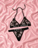 Conjunto de lencería de dos piezas confeccionado en encaje negro, compuesto por un soutiene triangular con tirantes halter y una bombacha tipo colaless. Presenta aplicaciones de letras brillantes con pedrería que forman la frase "FELIZ CUMPLE DISFRUTA".