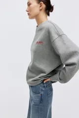 Buzo oversized de felpa color gris melange, con cuello redondo y hombros caídos. Cuenta con un pequeño bordado rojo con texto en el pecho y puños y bajo con ribete acanalado.
