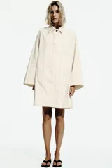 Trench de algodón color amarillo claro con patrón a cuadros sutiles de líneas oscuras. Tiene cuello con solapa, mangas largas, bolsillos delanteros y cierre frontal con botones ocultos bajo solapa.