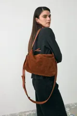Cartera mediana de gamuza color tostado, con diseño de doble asa y correa larga ajustable y desmontable. Cuenta con cierre superior y detalle de tiras laterales.